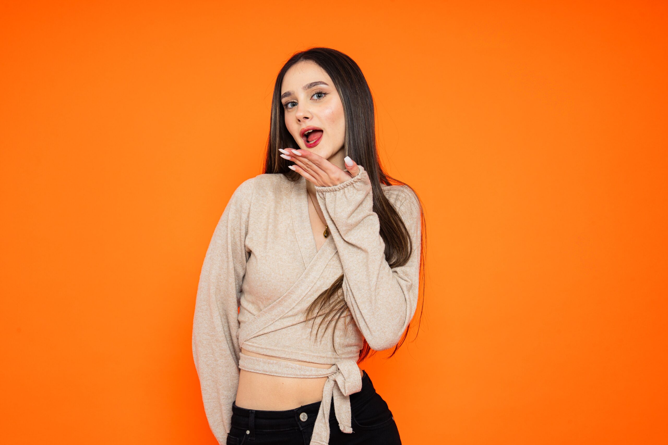 Stylish young woman in trendy outfit posing with playful expression on bright orange background.