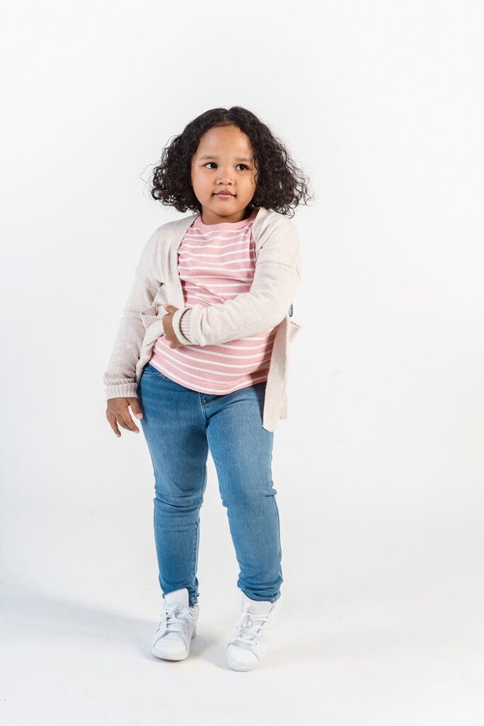 Full body of confident African American kid in jeans with cardigan and striped t shirt in studio