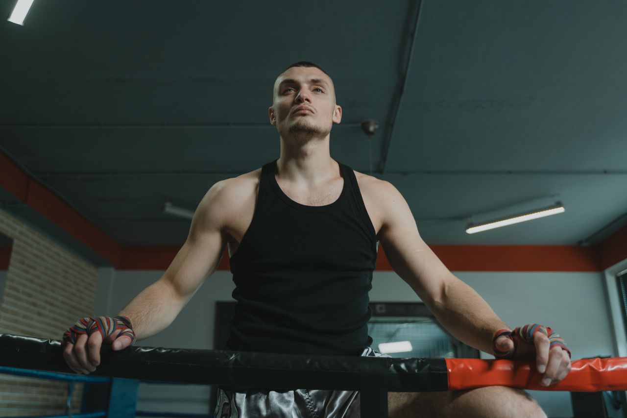 Muscular man in a boxing ring wearing black tank top and hand wraps, posing confidently.