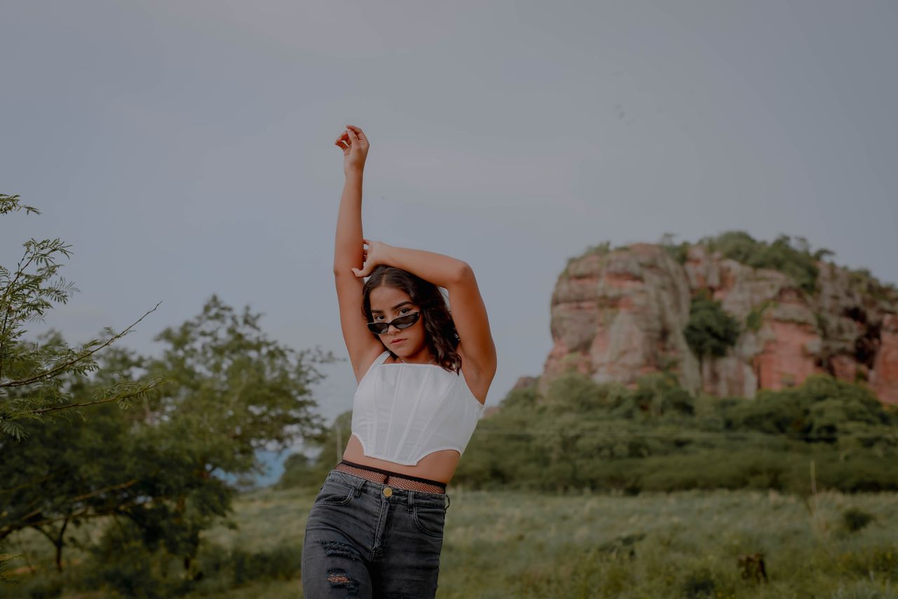 A stylish woman in sunglasses poses with arms raised against a natural rocky backdrop.