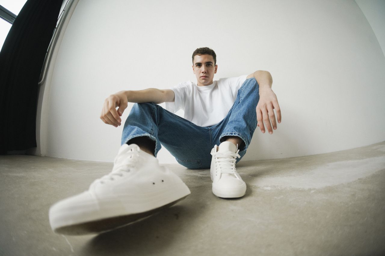 Young man in casual attire sitting in a minimalist studio, showcasing modern fashion style.